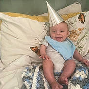 Charlie joined the competition — help win amazing prizes! baby, party_hat, blue_bib, crocheted_blanket, pillow, butterfly_pattern, bed, smile, happy, infant, clothing, foot, hand, headwear, fabric, indoor, person, cute, rest, cozy