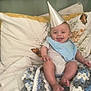 baby, party_hat, blue_bib, crocheted_blanket, pillow, butterfly_pattern, bed, smile, happy, infant, clothing, foot, hand, headwear, fabric, indoor, person, cute, rest, cozy