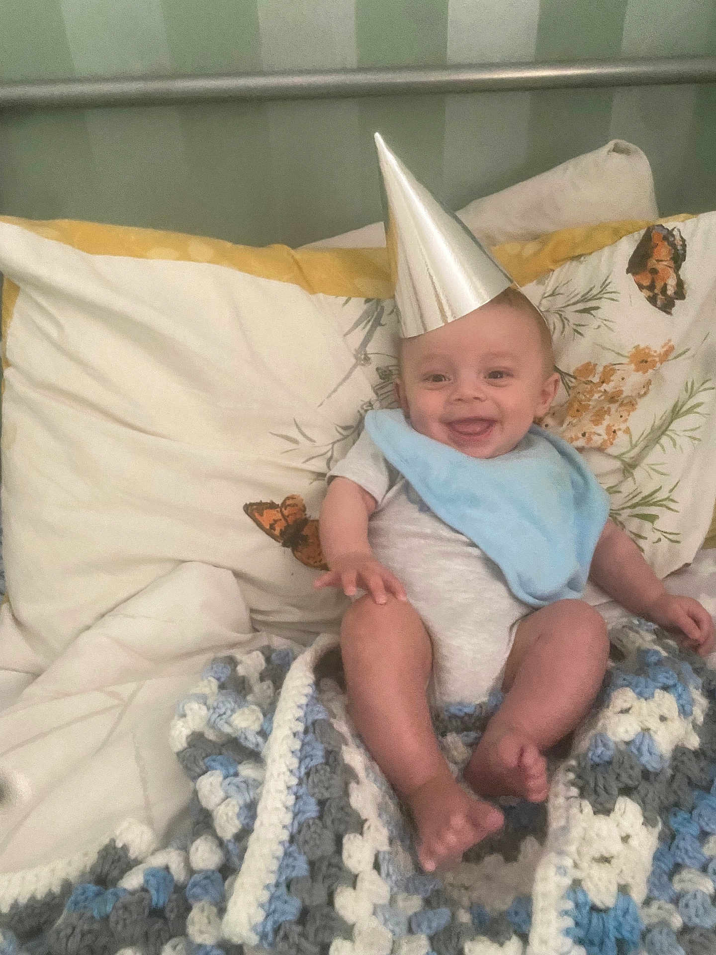Charlie joined the competition — help win amazing prizes! baby, party_hat, blue_bib, crocheted_blanket, pillow, butterfly_pattern, bed, smile, happy, infant, clothing, foot, hand, headwear, fabric, indoor, person, cute, rest, cozy