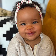 Thelma participe au concours pour gagner de l'argent avec cette photo : baby, child, smiling, headband, curly_hair, sweater, indoor, portrait, cute, cozy, face, person, seated, furniture, cushion, pattern, red_hearts, knitwear, expression, happy