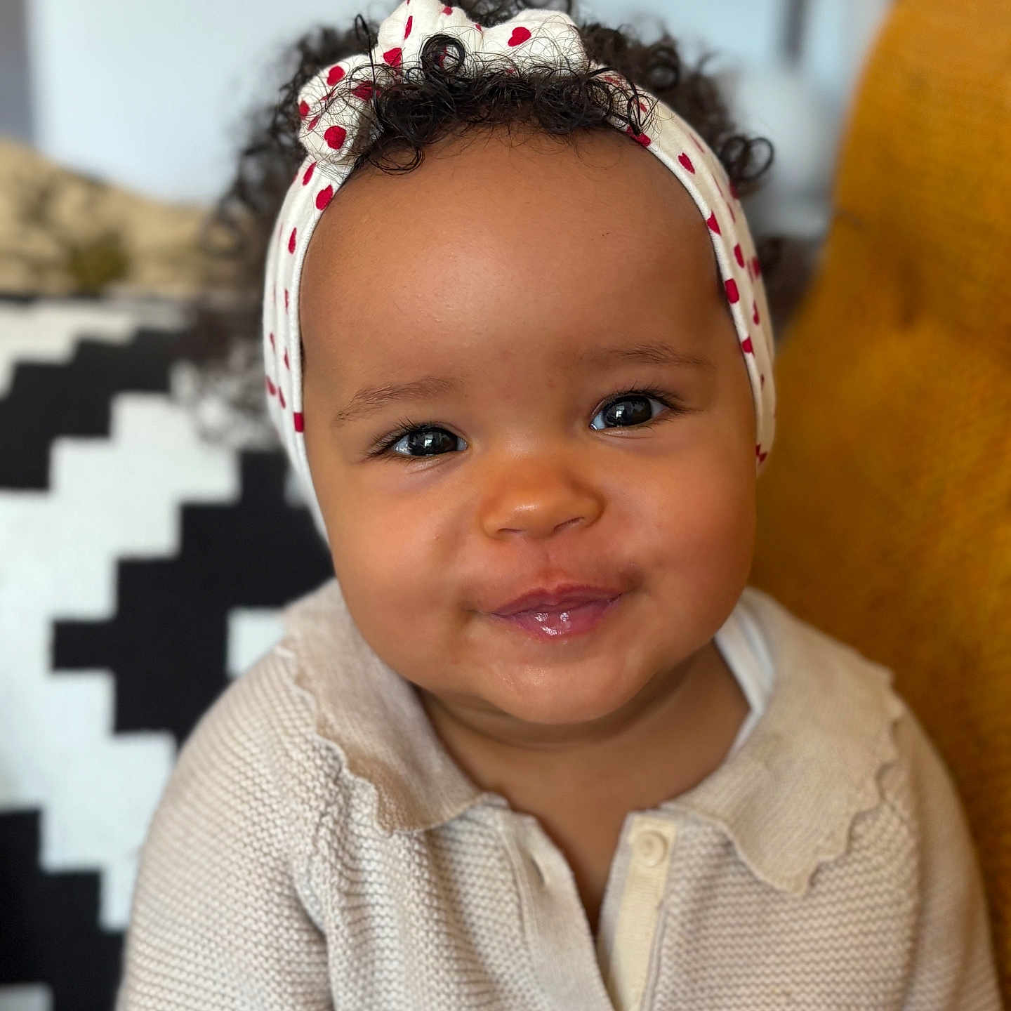 Thelma participe au concours pour gagner de l'argent avec cette photo : baby, child, cozy, curly_hair, cushion, cute, expression, face, furniture, happy, headband, indoor, knitwear, pattern, person, portrait, red_hearts, seated, smiling, sweater