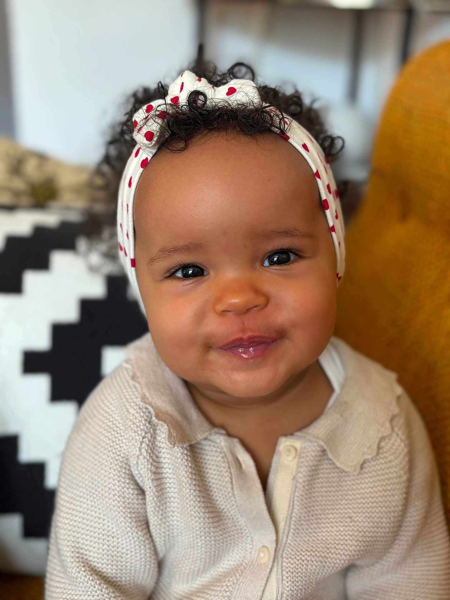 Thelma participe au concours pour gagner de l'argent avec cette photo : baby, child, smiling, headband, curly_hair, sweater, indoor, portrait, cute, cozy, face, person, seated, furniture, cushion, pattern, red_hearts, knitwear, expression, happy