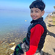 Amir a rejoint le concours — aidez-le/la à gagner de superbes lots ! child, boy, smiling, outdoor, lake, water, blue_sky, stone_ledge, casual_clothing, vest, red_shirt, nature, sunny_day, happy, portrait, person, park, rocks, reflection, daytime