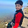 child, boy, smiling, outdoor, lake, water, blue_sky, stone_ledge, casual_clothing, vest, red_shirt, nature, sunny_day, happy, portrait, person, park, rocks, reflection, daytime