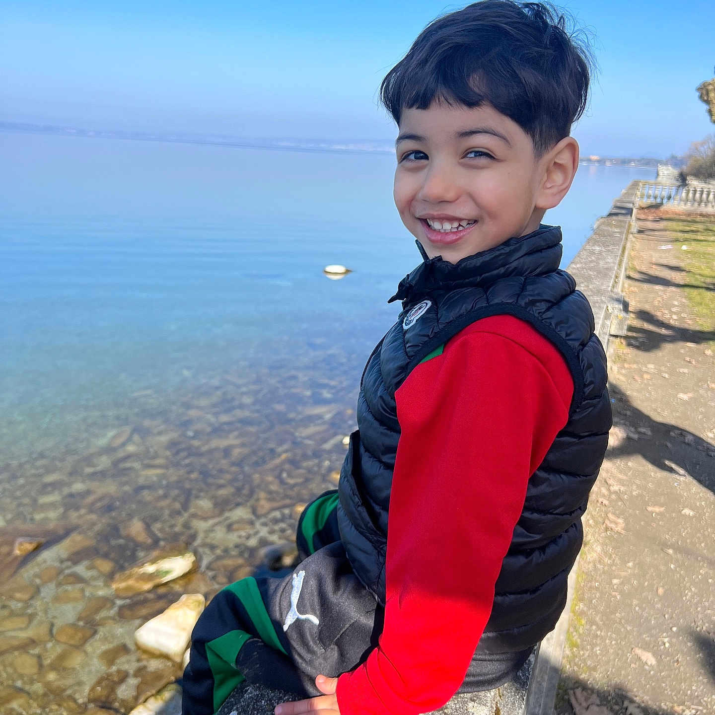 Amir a rejoint le concours — aidez-le/la à gagner de superbes lots ! child, boy, smiling, outdoor, lake, water, blue_sky, stone_ledge, casual_clothing, vest, red_shirt, nature, sunny_day, happy, portrait, person, park, rocks, reflection, daytime