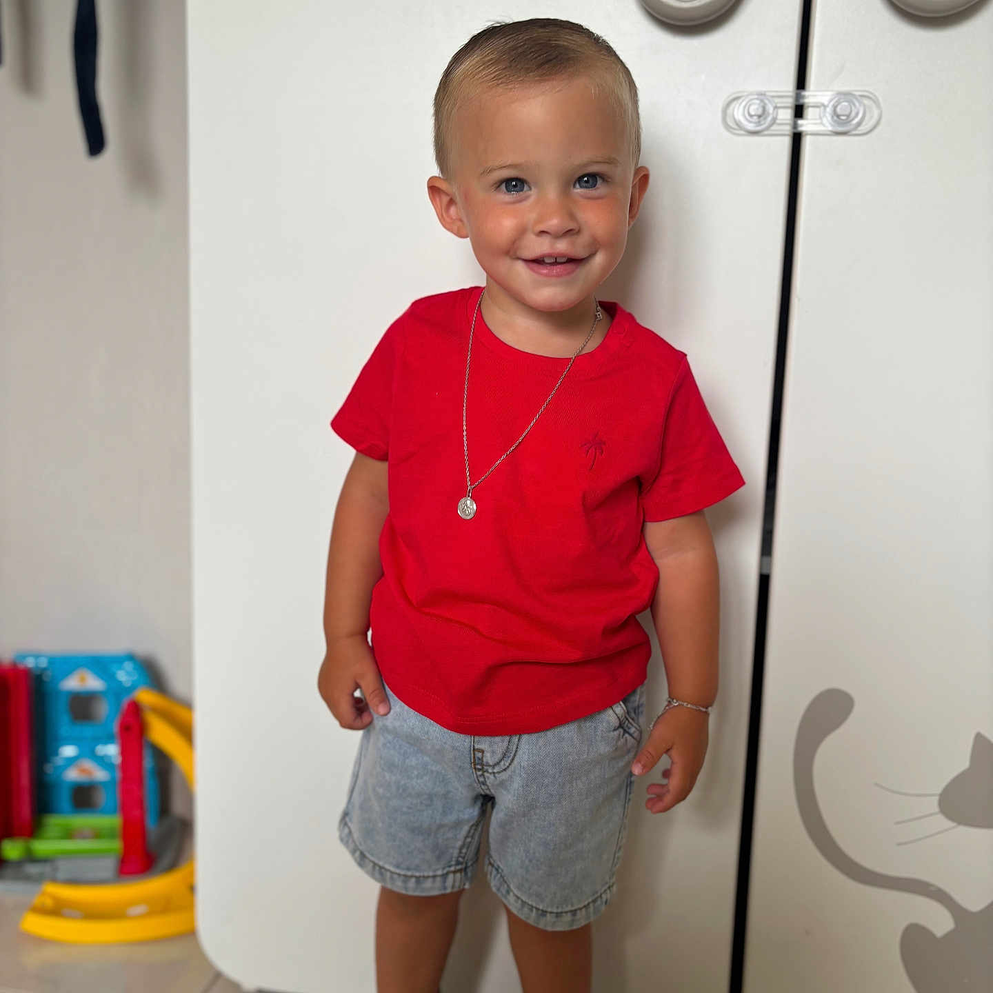 Lynaël a rejoint le concours — aidez-le/la à gagner de superbes lots ! bracelet, cabinet, casual_clothing, child, cute, denim_shorts, floor_tiles, happy, home, indoor, necklace, playroom, portrait, red_tshirt, smiling, sneakers, standing, toddler, toys, young_child