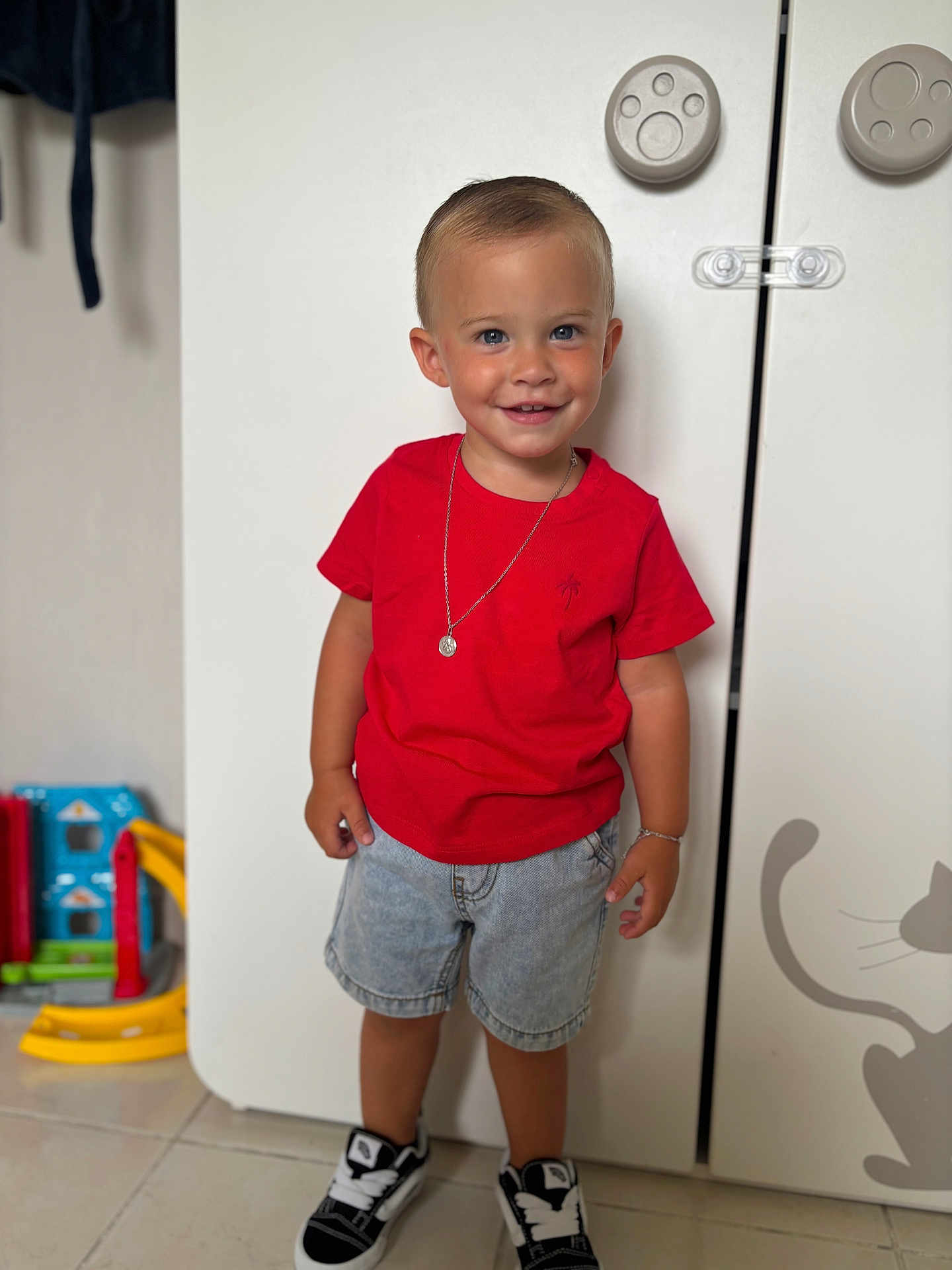 Lynaël a rejoint le concours — aidez-le/la à gagner de superbes lots ! bracelet, cabinet, casual_clothing, child, cute, denim_shorts, floor_tiles, happy, home, indoor, necklace, playroom, portrait, red_tshirt, smiling, sneakers, standing, toddler, toys, young_child