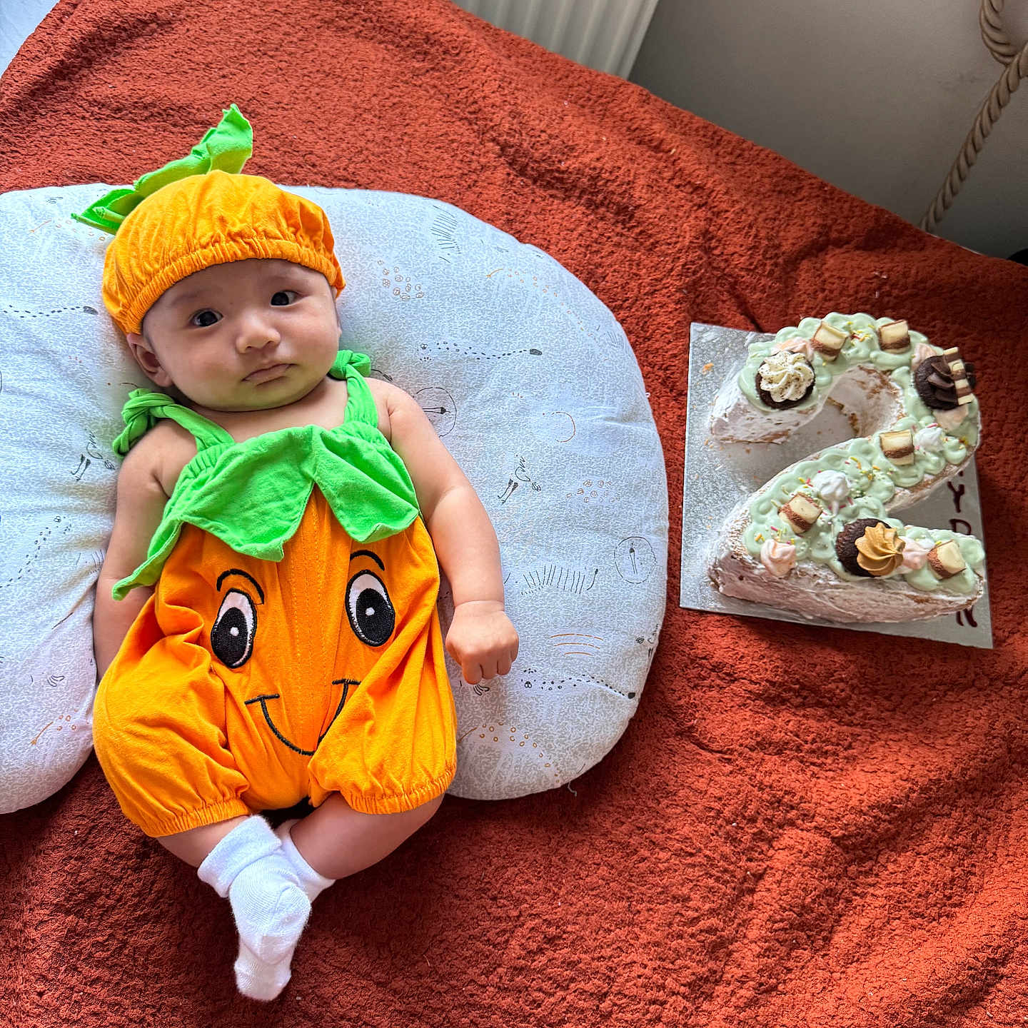 Rhayden joined the competition — help win amazing prizes! baby, blanket, cake, celebration, child, cute, decorated_cake, festive, food, green, indoor, infant, laying_down, number_two, orange, pillow, portrait, pumpkin_costume, smile, socks