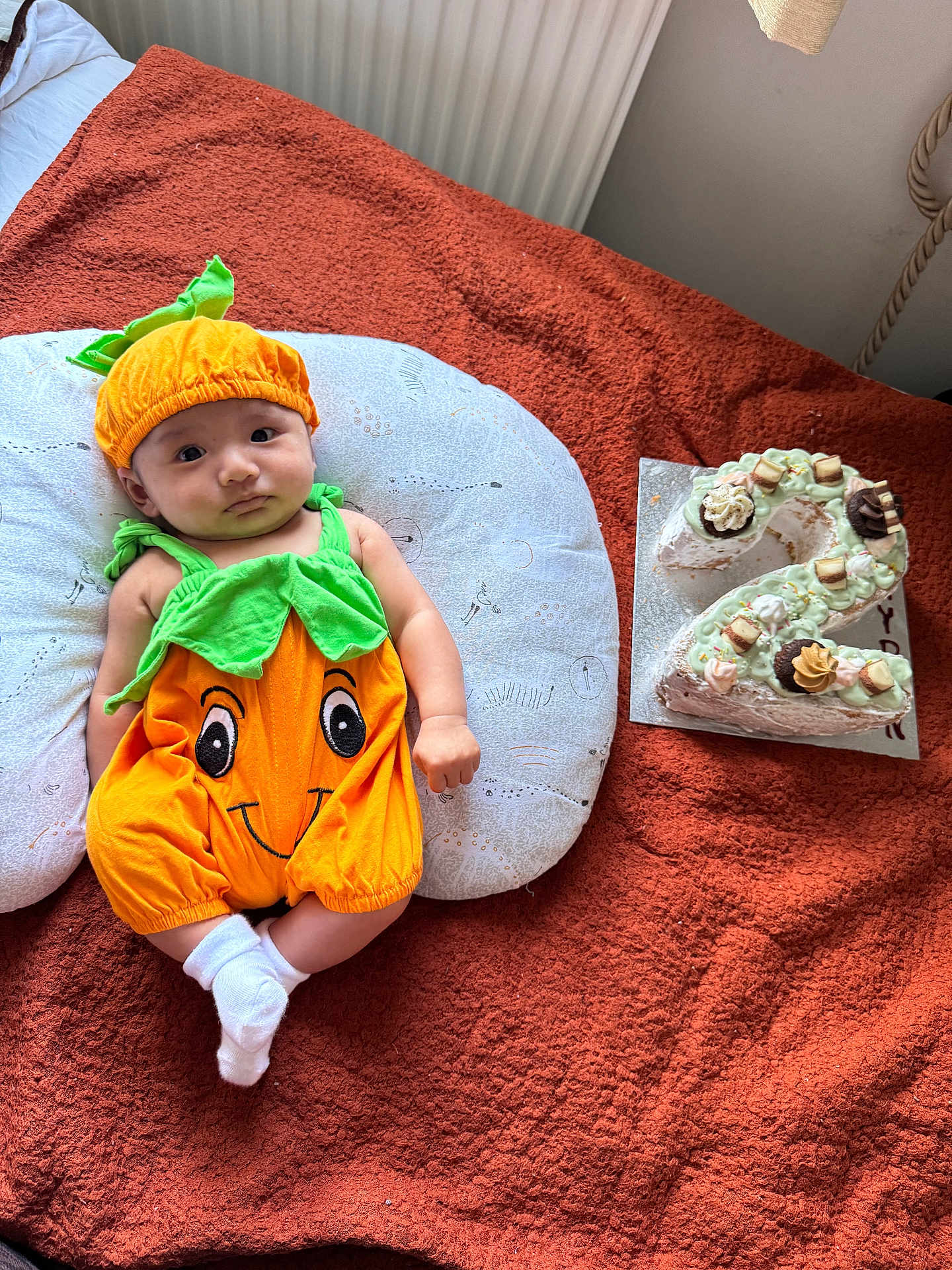 Rhayden joined the competition — help win amazing prizes! baby, infant, pumpkin_costume, orange, green, pillow, blanket, cake, number_two, celebration, smile, cute, socks, indoor, portrait, child, festive, food, decorated_cake, laying_down