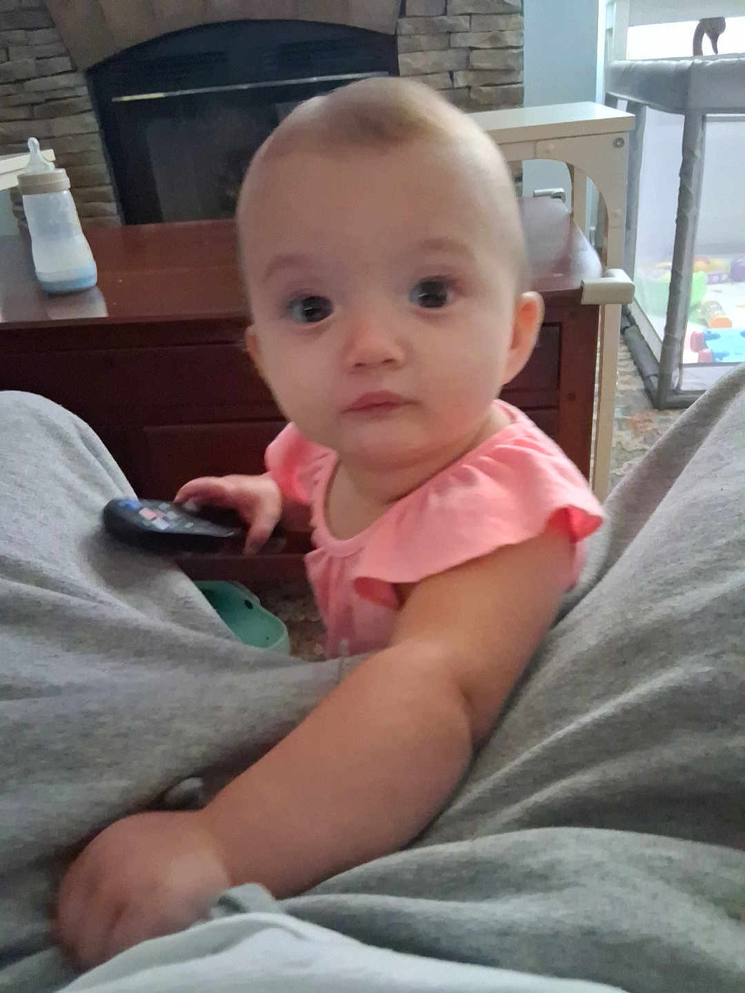 Autumn is registered to the contest to win money with this photo: arm, baby, bottle, carpet, child, couch, curious, face, fireplace, furniture, home, indoor, infant, living_room, person, pink_clothing, remote_control, sitting, table, toy