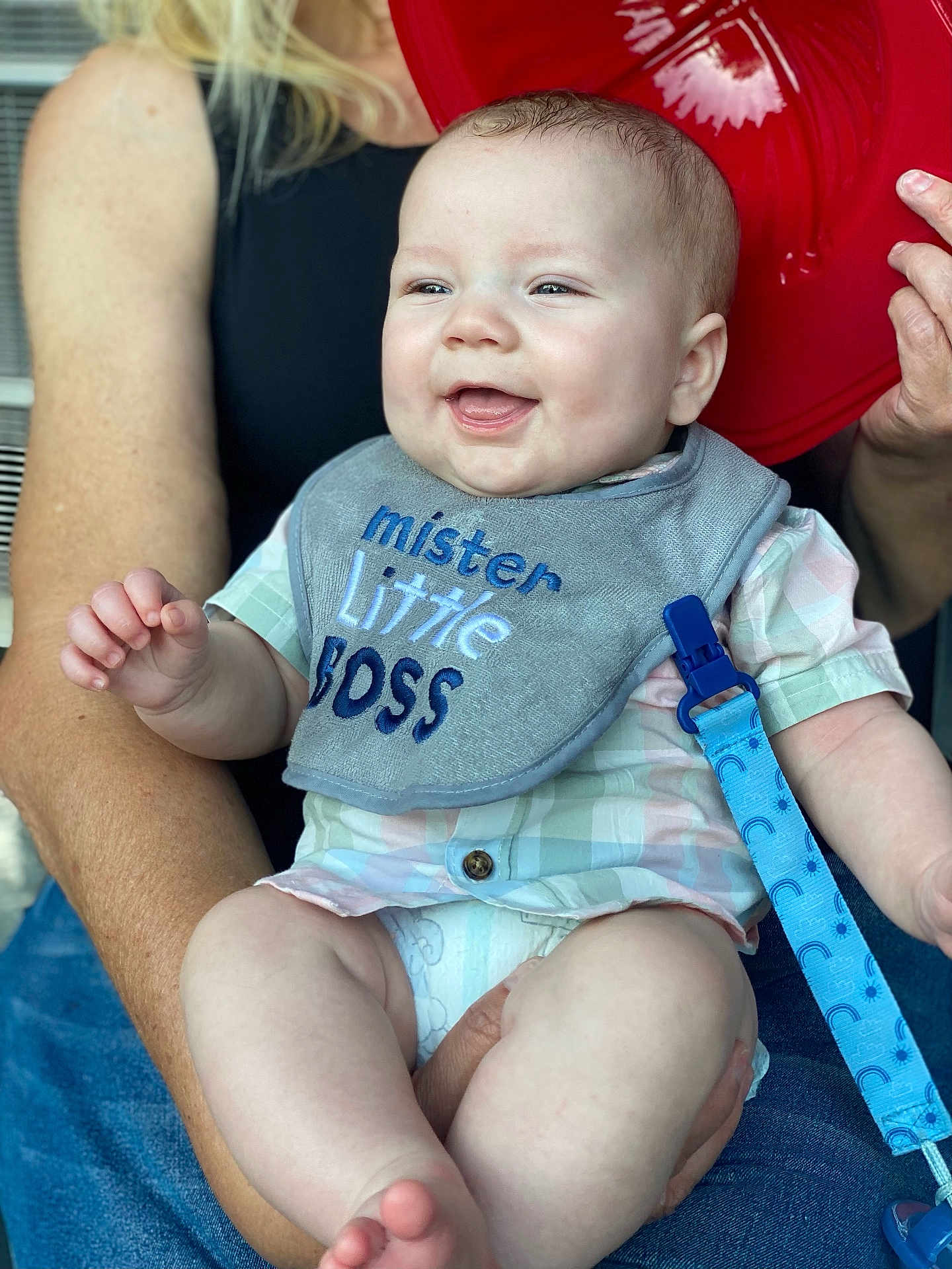 Phoenix is registered to the contest to win money with this photo: baby, infant, smiling, bib, clothing, person, adult, hand, face, skin, arm, red_object, blue_clip, diaper, jeans, sitting, happy, casual, indoor, portrait