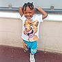 child, smiling, braids, cartoon_clothing, shorts, tshirt, sneakers, outdoor, wall, pavement, happy, standing, casual, playful, portrait, person, young, fashion, cute, daylight