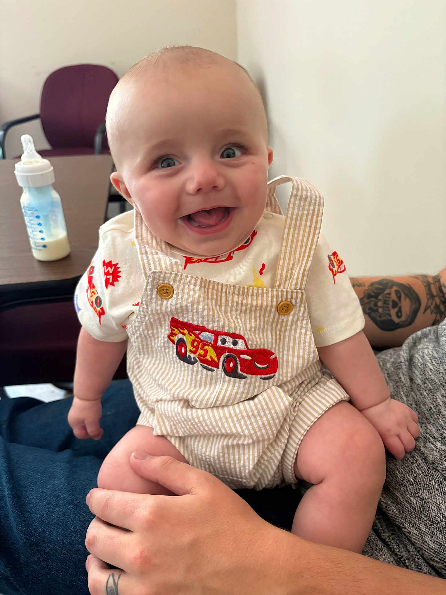 John Lee is registered to the contest to win money with this photo: baby, smiling, overalls, race_car, tattoo, hand, bottle, chair, table, indoor, cute, happy, child, person, clothing, skin, face, sitting, holding, casual