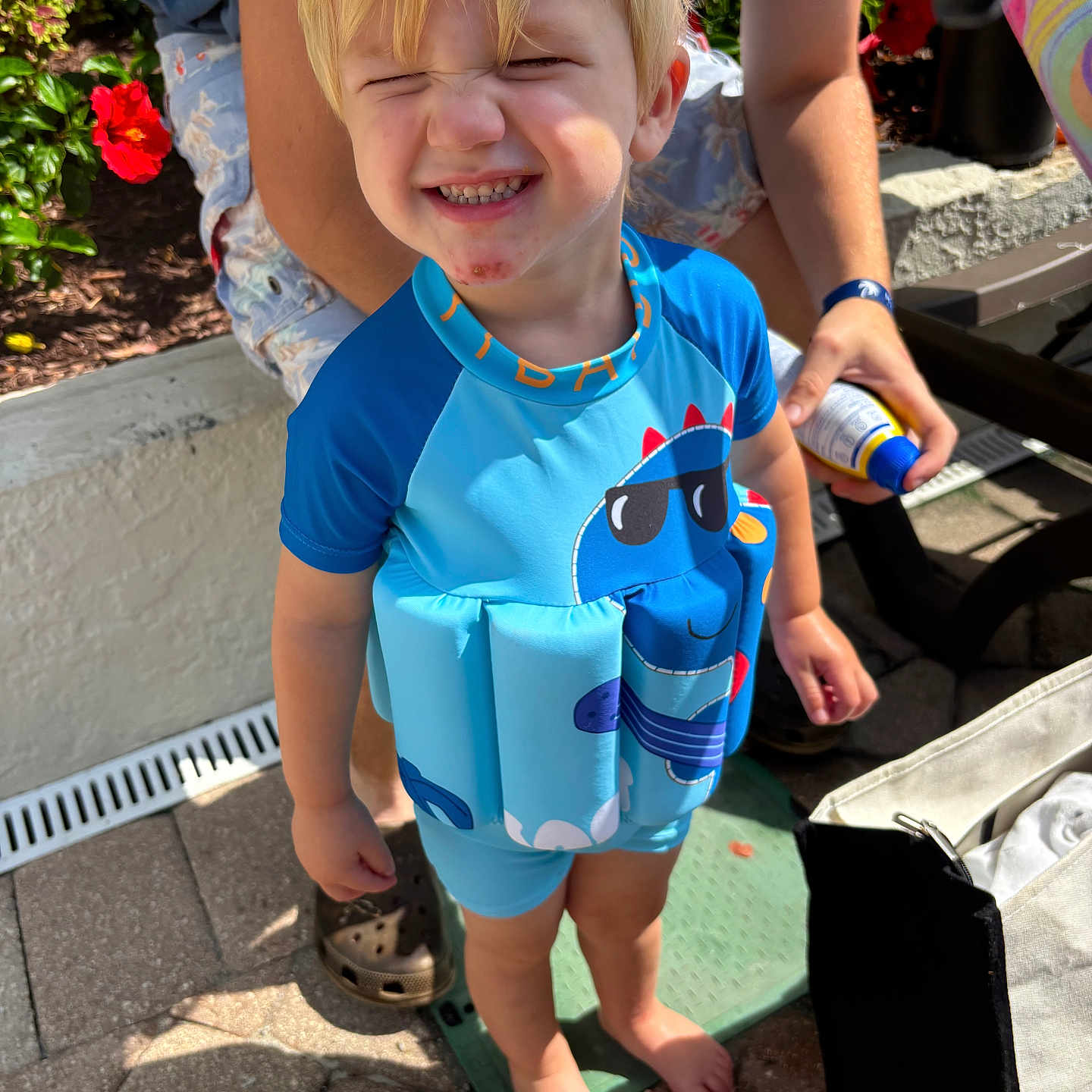 Thomas Brock joined the competition — help win amazing prizes! adult_hand, barefoot, blue_clothing, casual, child, daytime, dinosaur_print, flotation_vest, flowers, greenery, happy, outdoor, pavement, person, playful, smiling, summer, sunlight, sunscreen, toddler