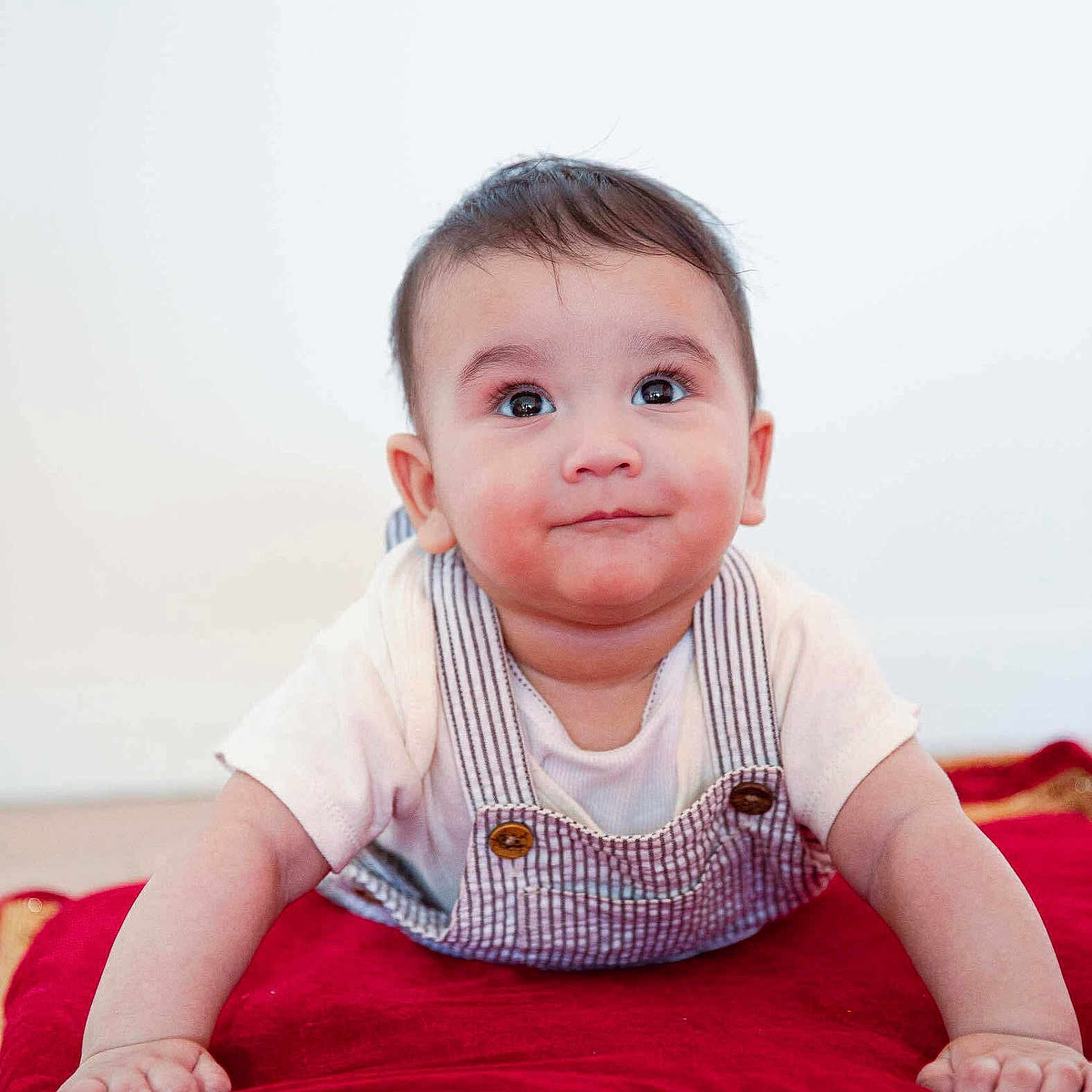 Arvik is registered to the contest to win money with this photo: baby, child, cute, expression, eyes, face, hair, hands, indoor, infant, lying_down, overalls, person, plush, portrait, red_cushion, shirt, skin, smile, young