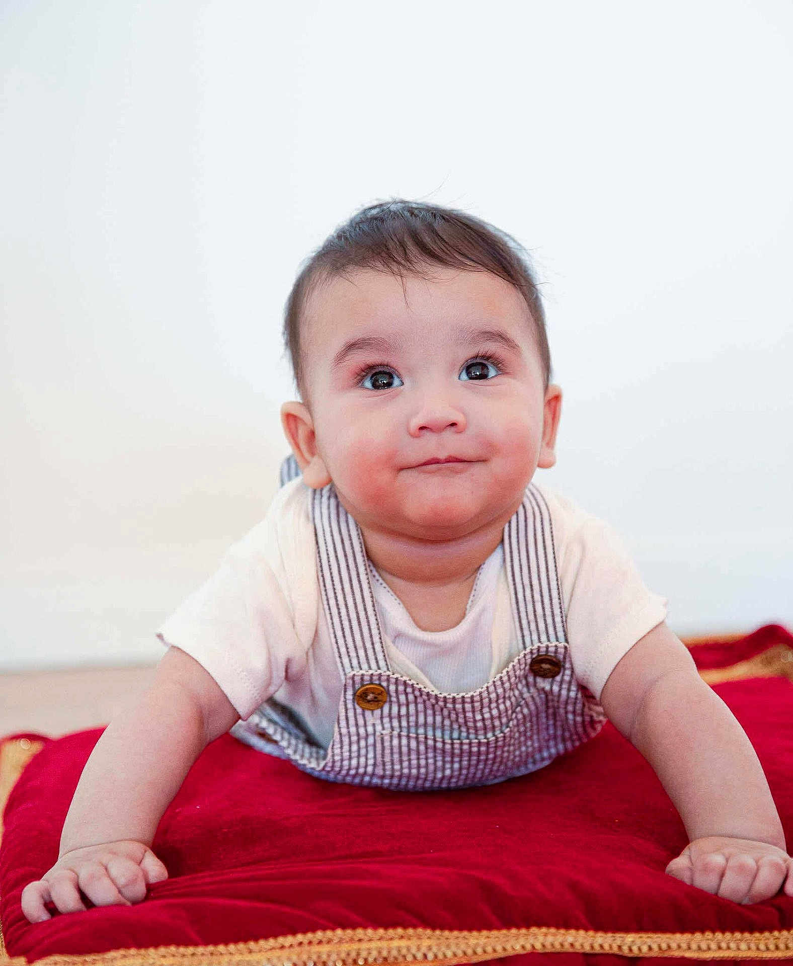 Arvik is registered to the contest to win money with this photo: baby, infant, child, face, eyes, smile, overalls, shirt, red_cushion, plush, cute, portrait, indoor, person, hands, expression, skin, hair, young, lying_down
