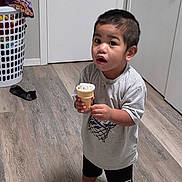 Aron is registered to the contest to win money with this photo: child, toddler, boy, ice_cream, cone, dessert, surprised, curious, standing, barefoot, indoors, kitchen, wood_floor, laundry_basket, clothing, t_shirt, shorts, portrait, face, smile