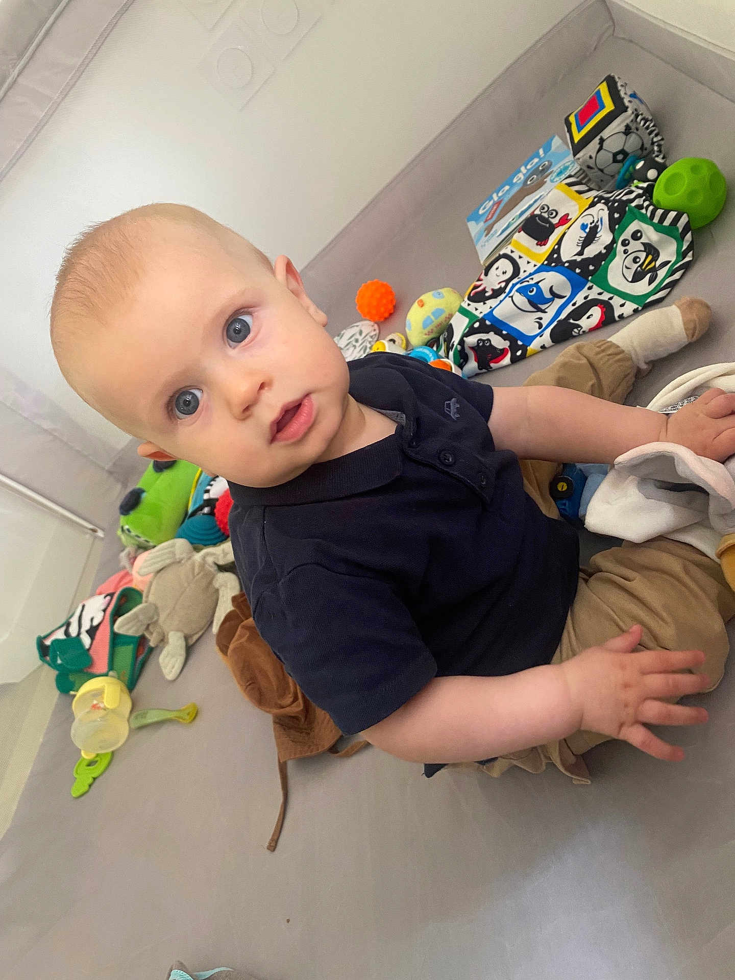 Théo a rejoint le concours — aidez-le/la à gagner de superbes lots ! baby, child, indoor, toys, playmat, blue_eyes, polo_shirt, tan_pants, blankets, plush_toys, colorful, curious, sitting, infant, hand, face, expression, soft_surface, playtime, young