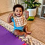 baby, bib, blue_onesie, cabinet, carpet, child, curly_hair, door, fist_raised, floor, happy, home, indoor, person, plant, play_mat, potted_plant, rug, smiling, toy_block