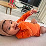 baby, infant, child, orange_onesie, head_wrap, lying_down, smiling, indoors, living_room, table, television, curtains, floor, toy, furniture, decorations, holiday, cute, portrait, playful
