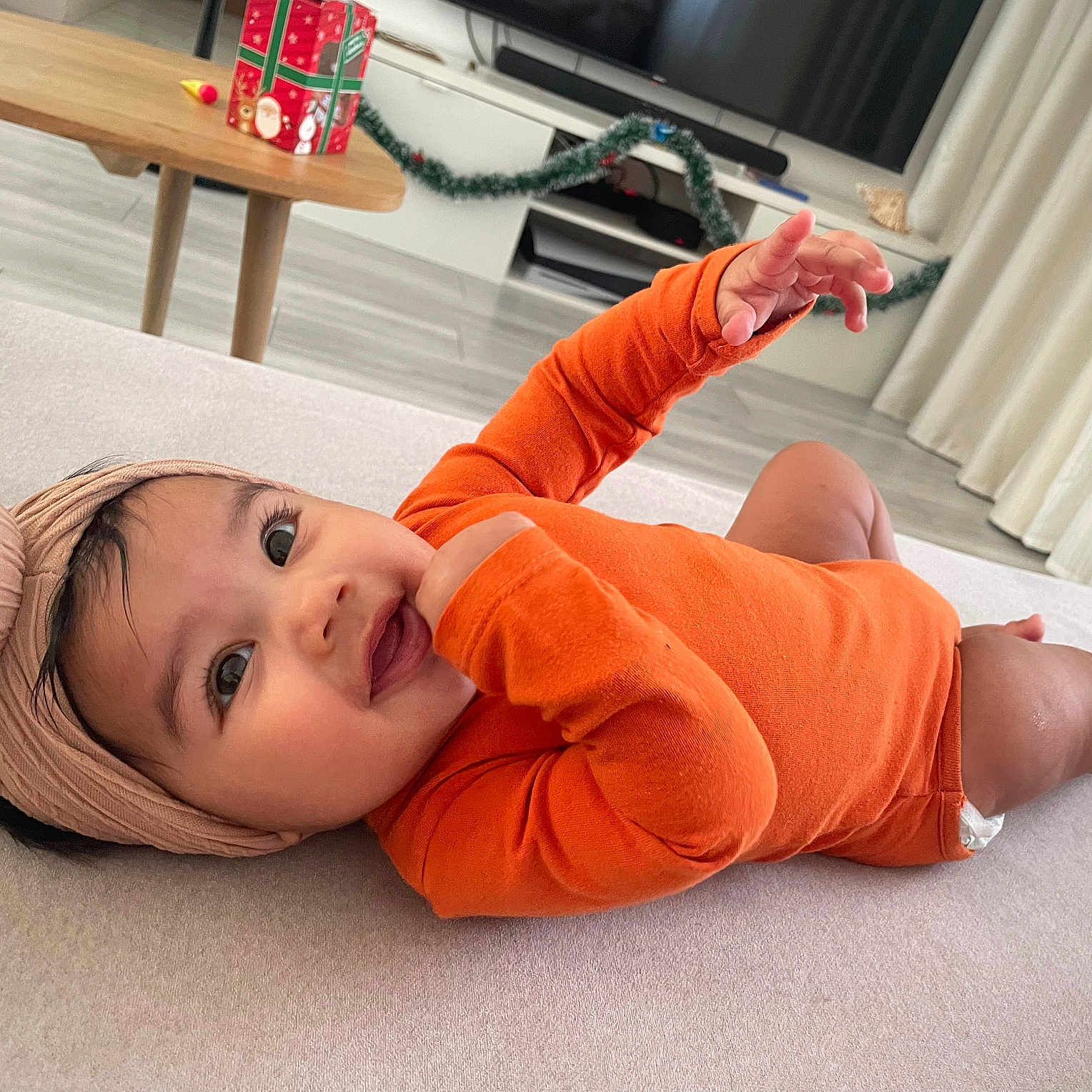 Alya participe au concours pour gagner de l'argent avec cette photo : baby, child, curtains, cute, decorations, floor, furniture, head_wrap, holiday, indoors, infant, living_room, lying_down, orange_onesie, playful, portrait, smiling, table, television, toy