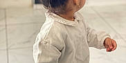 Alya participe au concours pour gagner de l'argent avec cette photo : back_view, beige_pants, blurred_background, casual_clothing, child, cute, earring, furniture, hair_bun, holding, home, indoor, living_room, microphone, person, standing, tile_floor, toddler, toy, white_sweater