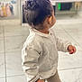 back_view, beige_pants, blurred_background, casual_clothing, child, cute, earring, furniture, hair_bun, holding, home, indoor, living_room, microphone, person, standing, tile_floor, toddler, toy, white_sweater