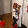 casual_clothing, child, curious, door, girl, hair_beads, hand_on_door, home, indoors, memory, portrait, red_boots, room, screen, smartphone_screenshot, social_media, standing, white_tshirt, wood_floor, young_child