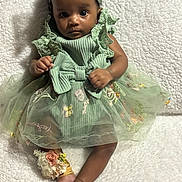 Yanni joined the competition — help win amazing prizes! baby, child, green_dress, headband, floral_embroidery, anklet, barefoot, curly_hair, sitting, blanket, textured_blanket, indoor, portrait, cute, infant, clothing, footwear, accessory, person, young_child