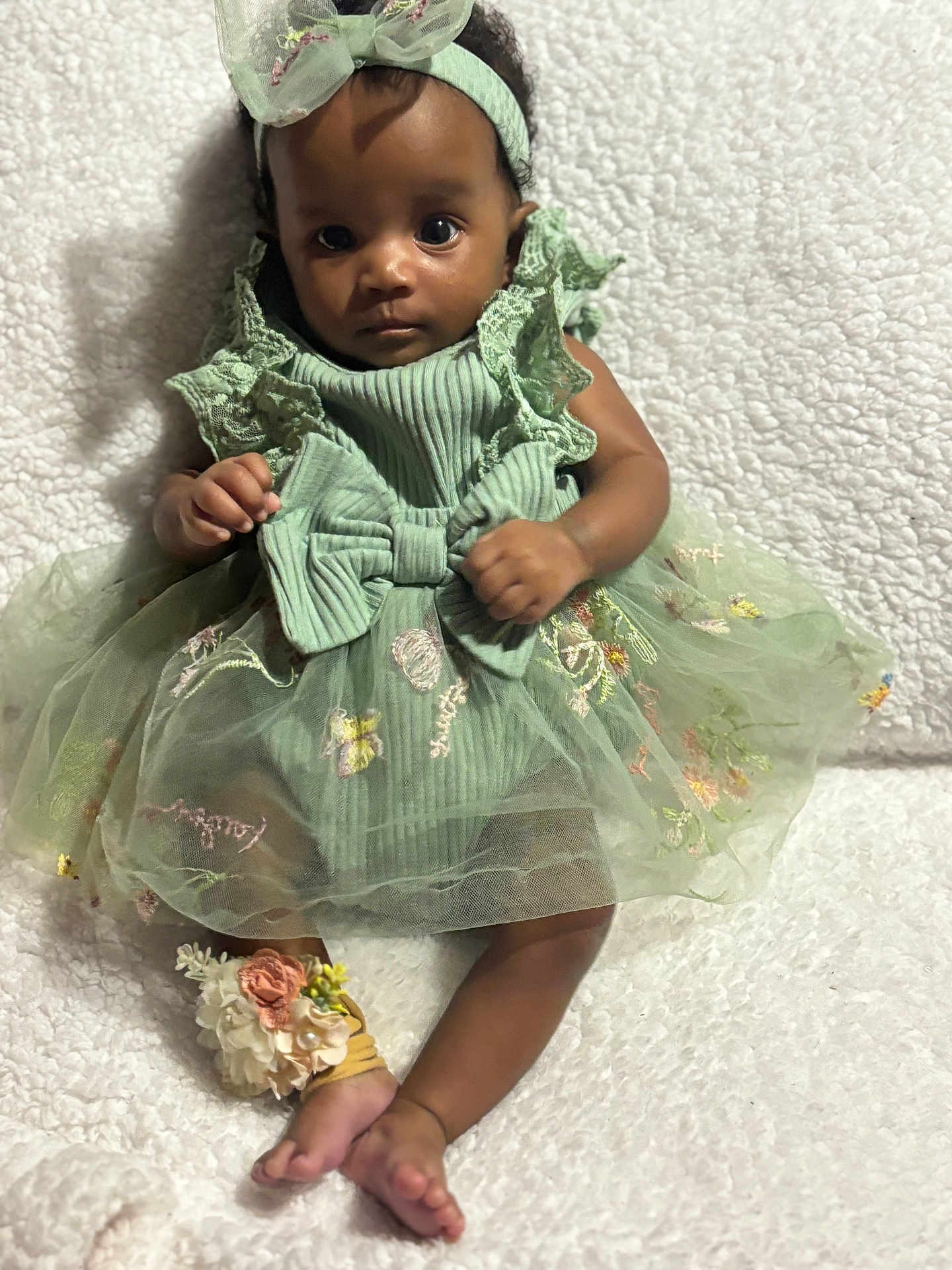 Yanni joined the competition — help win amazing prizes! baby, child, green_dress, headband, floral_embroidery, anklet, barefoot, curly_hair, sitting, blanket, textured_blanket, indoor, portrait, cute, infant, clothing, footwear, accessory, person, young_child
