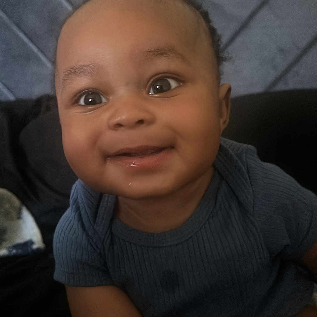 Micah joined the competition — help win amazing prizes! baby, smiling, child, face, portrait, blue_clothing, happy, cute, indoors, seated, head, skin, person, infant, young_child, expression, hands, background, textile, quilting