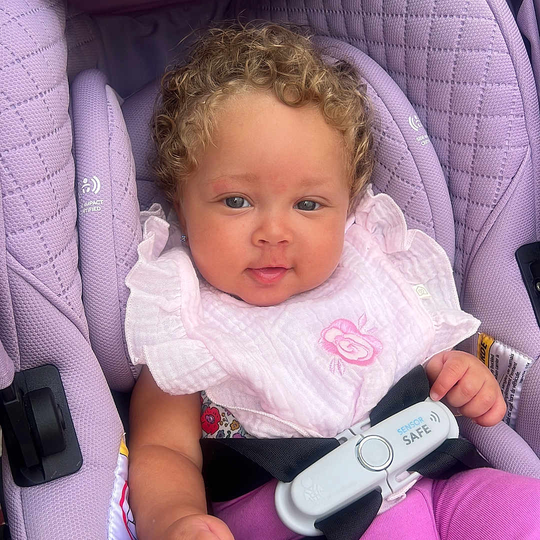 Lia is registered to the contest to win money with this photo: baby, child, infant, car_seat, lavender, pink_clothing, ruffled_shirt, curly_hair, portrait, sitting, harness, safety_buckle, vehicle_interior, indoors, face, eyes, hands, fashion, adorable, photograph