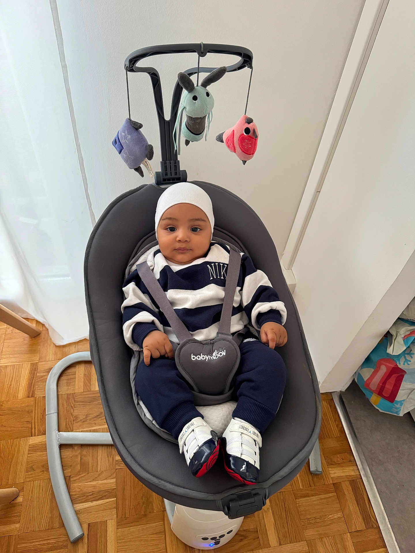 Adam a rejoint le concours — aidez-le/la à gagner de superbes lots ! baby, infant, baby_rocker, toy_mobile, striped_clothing, nike, white_hat, baby_shoes, indoor, wooden_floor, curtain, gray_furniture, child, person, cute, seated, playful, soft_toys, calm, portrait