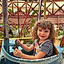 child, curly_hair, smile, amusement_ride, steering_wheel, vintage, blue_car, outdoor, daylight, metal, rust, plants, greenery, carnival, playful, happy, person, seat, striped_shirt, overalls