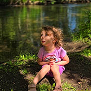 Brylee is registered to the contest to win money with this photo: child, curly_hair, pink_shirt, sandals, sitting, forest, river, greenery, sunlight, nature, outdoor, grass, dirt, daylight, portrait, young_child, casual_clothing, peaceful, curious, environment