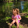 casual_clothing, child, curious, curly_hair, daylight, dirt, environment, forest, grass, greenery, nature, outdoor, peaceful, pink_shirt, portrait, river, sandals, sitting, sunlight, young_child