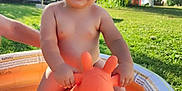 Sacha a rejoint le concours — aidez-le/la à gagner de superbes lots ! bare_chest, child, daylight, garden, giraffe_toy, grass, greenery, happy, inflatable_pool, orange_toy, outdoor, person, playtime, smiling, summer, sunny, toddler, toy, trees, water
