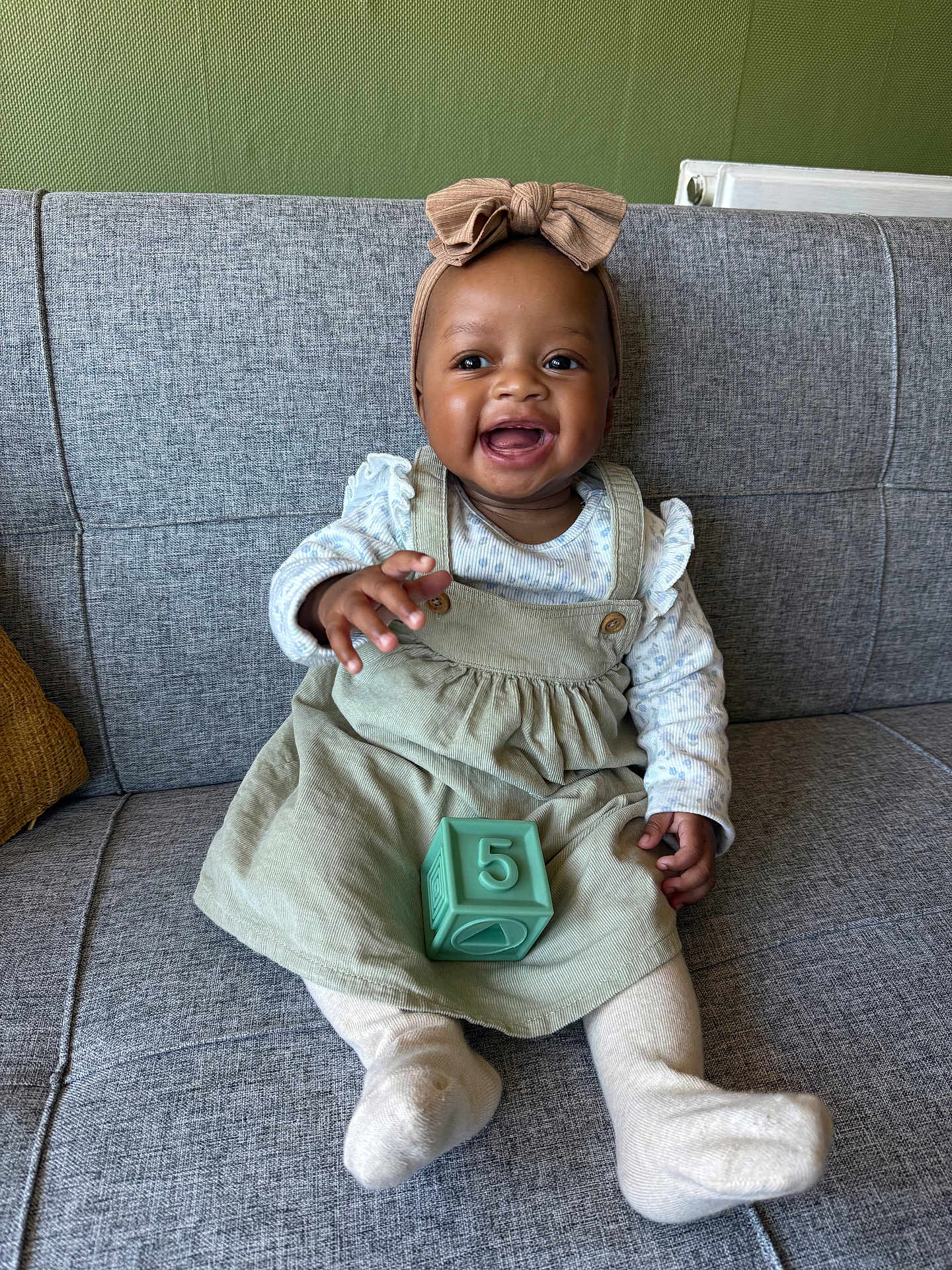 Charis joined the competition — help win amazing prizes! baby, child, smiling, sitting, couch, gray_couch, green_dress, headband, bow, toy_block, number_5, happy, indoor, person, long_sleeve, tights, radiator, wall, furniture, cute