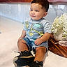 toddler, child, boy, sitting, smiling, portrait, indoor, kitchen, countertop, wooden_cabinets, flowers, bouquet, white_flowers, sneakers, black_shoes, denim_shorts, graphic_shirt, necklace, cross_pendant, home_decor