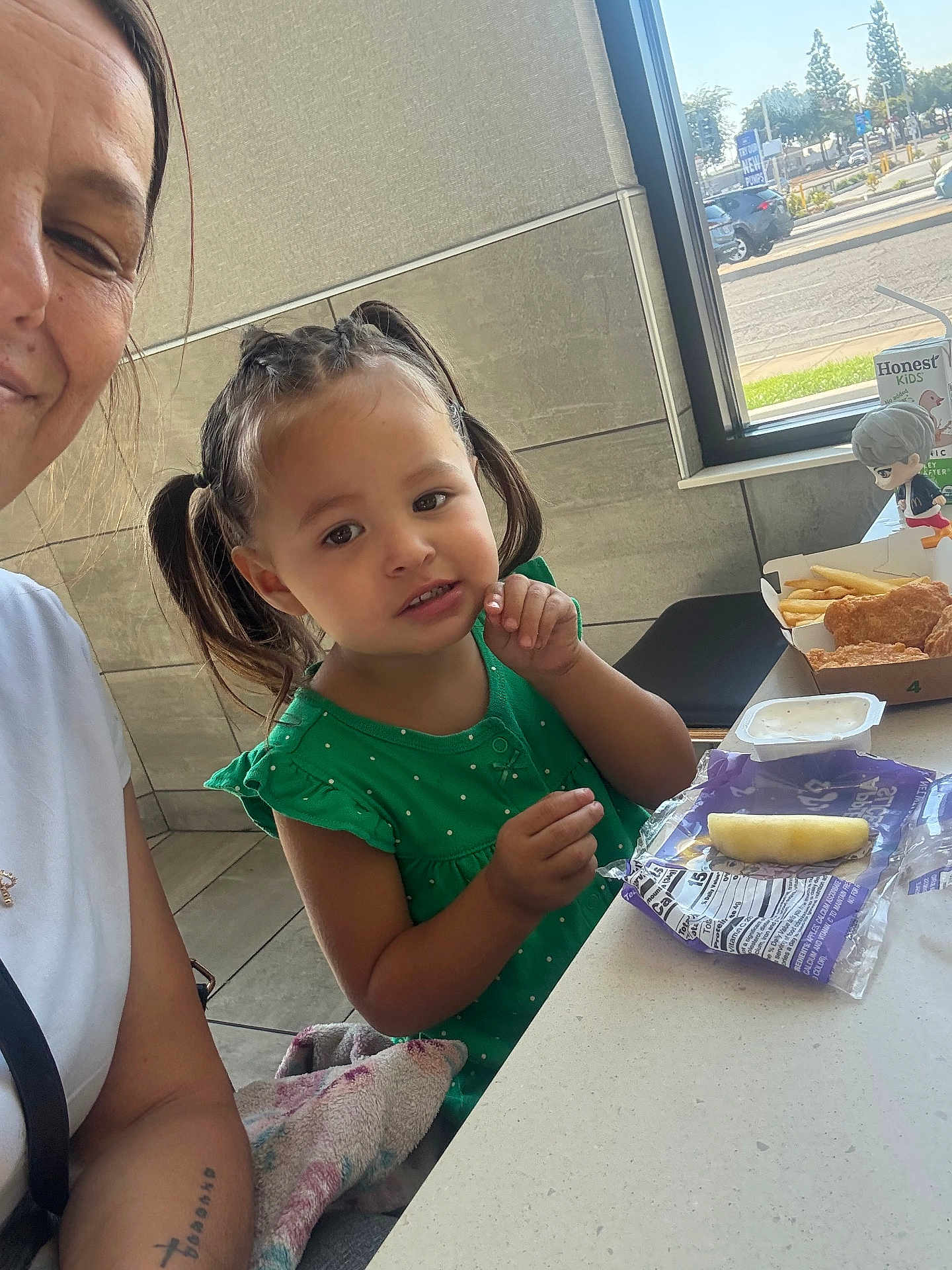Kayleah is registered to the contest to win money with this photo: child, girl, woman, green_dress, pigtails, table, food, fries, chicken_nuggets, dipping_sauce, snack_pack, figurine, window, parking_lot, indoor, curious, person, face, casual, restaurant