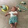 baby, infant, smiling, blue_shorts, white_shirt, bear_print, cake, teddy_bear, decorative_hoop, knitted_yarn, pom_poms, name_sign, textile, blanket, floor, cute, child, portrait, indoors, celebration