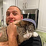 man, cat, hat, kitchen, cabinet, microwave, countertop, appliance, indoor, pet, holding, expression, face, arm, clothing, white_cabinet, modern, human, animal, funny