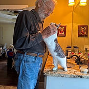 Timber is registered to the contest to win money with this photo: elderly_man, cat, bathroom, mirror, countertop, lamp, portrait, jeans, glasses, beard, petting, indoor, home, warm_lighting, white_cat, gray_spots, gentle, affection, casual_clothing, reflection