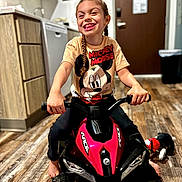 Mckenzie is registered to the contest to win money with this photo: child, smiling, toy_vehicle, indoor, kitchen, braided_hair, mickey_mouse, tshirt, floor, wood_flooring, playful, happy, person, footwearless, plastic_toy, small_vehicle, plush_toy, cabinet, door, handlebars