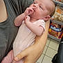 baby, infant, person, holding, onesie, hand, face, nose_picking, adult, gray_shirt, store, aisle, shelf, pet_food, tile_floor, shopping, care, cradle, curious, cute
