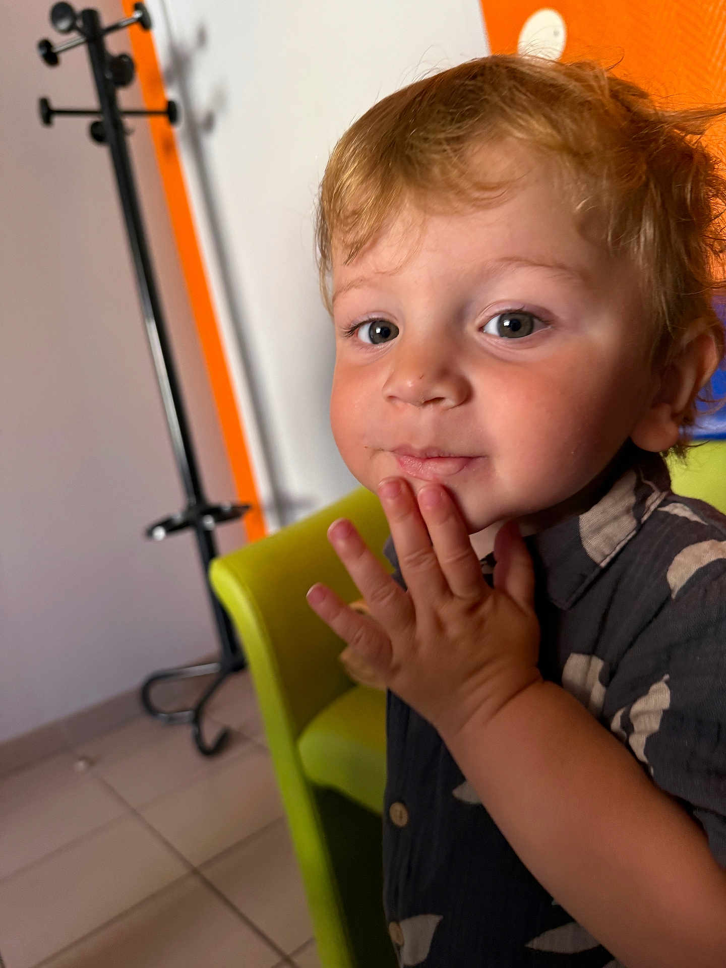 Kurtys a rejoint le concours — aidez-le/la à gagner de superbes lots ! toddler, child, portrait, indoor, curly_hair, blond_hair, green_chair, hand_on_face, cute, casual_clothing, buttoned_shirt, floor_tiles, coat_rack, orange_wall, white_wall, expression, closeup, person, young_child, playful