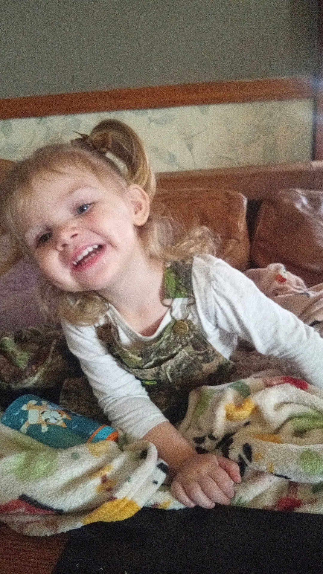 Aurora is registered to the contest to win money with this photo: child, girl, smiling, blonde_hair, pigtails, blanket, couch, indoor, happy, water_bottle, long_sleeve_shirt, overalls, home, playful, portrait, young, cute, cozy, furniture, relaxed