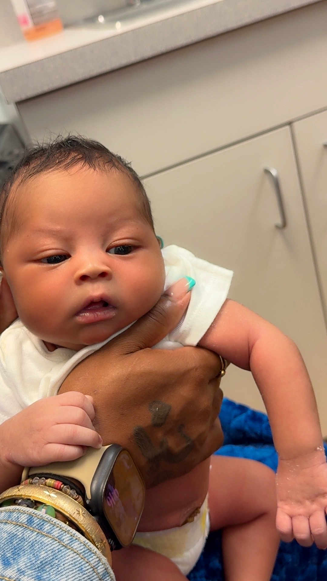 Racari Thompson joined the competition — help win amazing prizes! baby, infant, hand, tattoo, bracelet, smartwatch, diaper, white_clothing, skin, face, arm, fingers, cabinet, blue_blanket, indoor, person, holding, closeup, nursery, care