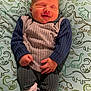 baby, smiling, infant, dinosaur_pattern, pillow, blue_clothing, grey_clothing, textured_fabric, lying_down, cute, small_hands, closed_eyes, happy, child, newborn, patterned_socks, indoor, cozy, soft_fabric, person