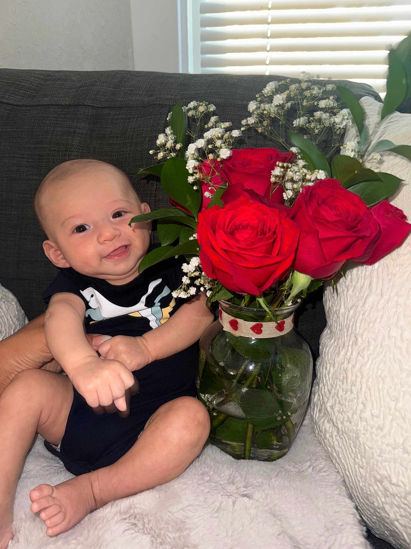 Silviano joined the competition — help win amazing prizes! baby, infant, smile, black_onesie, red_roses, flower_vase, bouquet, green_leaves, glass_vase, heart_ribbon, white_blanket, couch, window_blinds, indoor, happy, person, skin, hand, foot, plant